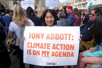 Joan, Asian woman, is standing in a crowd holding a large poster saying 'Tony Abbot: Climate action is on MY agenda'