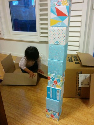 There is a colourful stack of tissue boxes. Behind the stack, there are two cardboard boxes. A toddler is sitting in one of them. Her face is not visible.