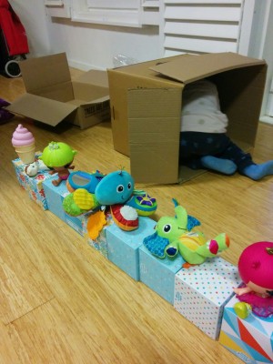 A range of colourful toys sit on top of a row of colourful tissue boxes. There are two boxes in the background. A toddler's legs and feet are sticking out of one of the boxes.