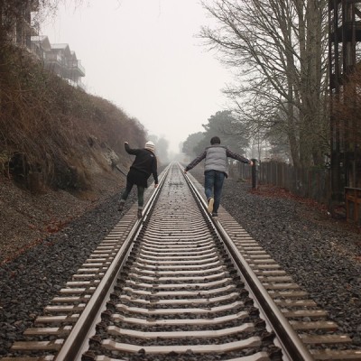 Two people standing on railway tracks next to each other. The tracks travel into the distance. Sky is gray and trees have lost their leaves.