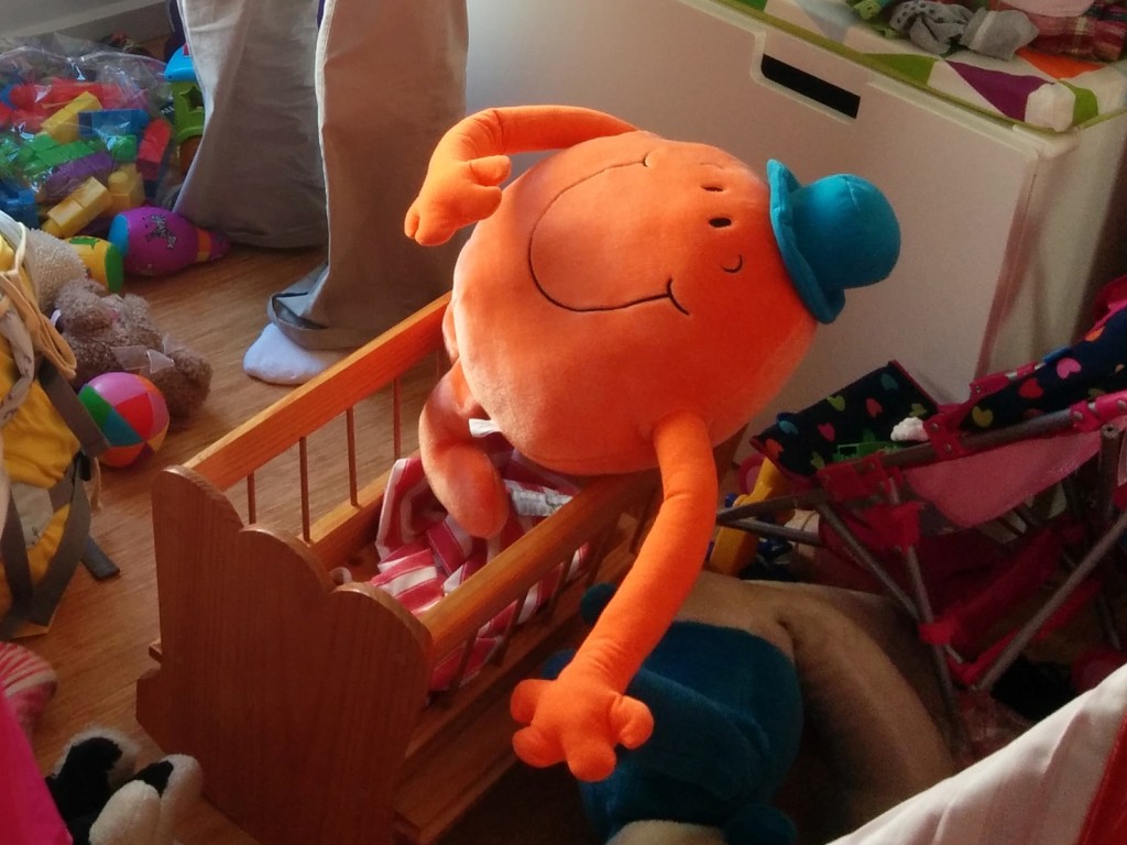 Mr Tickle, a bright orange stuffed toy with long arms and a blue hat, is sitting in a toy cradle.